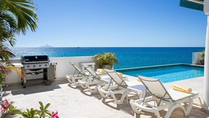 Zona de piscina con sillones de descanso y una parrilla, con vista al mar azul y cristalino.