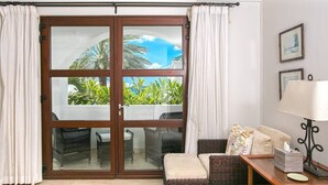 Habitación con puerta de vidrio corrediza que da a un balcón con mobiliario exterior y vegetación. Incluye un sofá de mimbre, una mesita con lámpara y pañuelos.