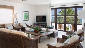 Un salón amplio con muebles de mimbre, un televisor de pantalla plana y un gran ventanal con vistas a palmeras.