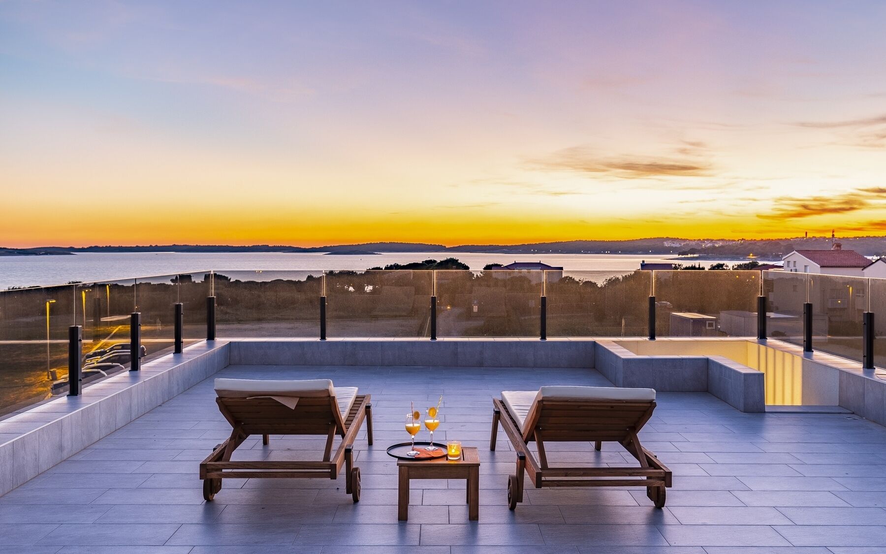 Une terrasse sur le toit, avec deux fauteuils en bois, une petite table avec des boissons et une vue sur le coucher de soleil sur l’eau.