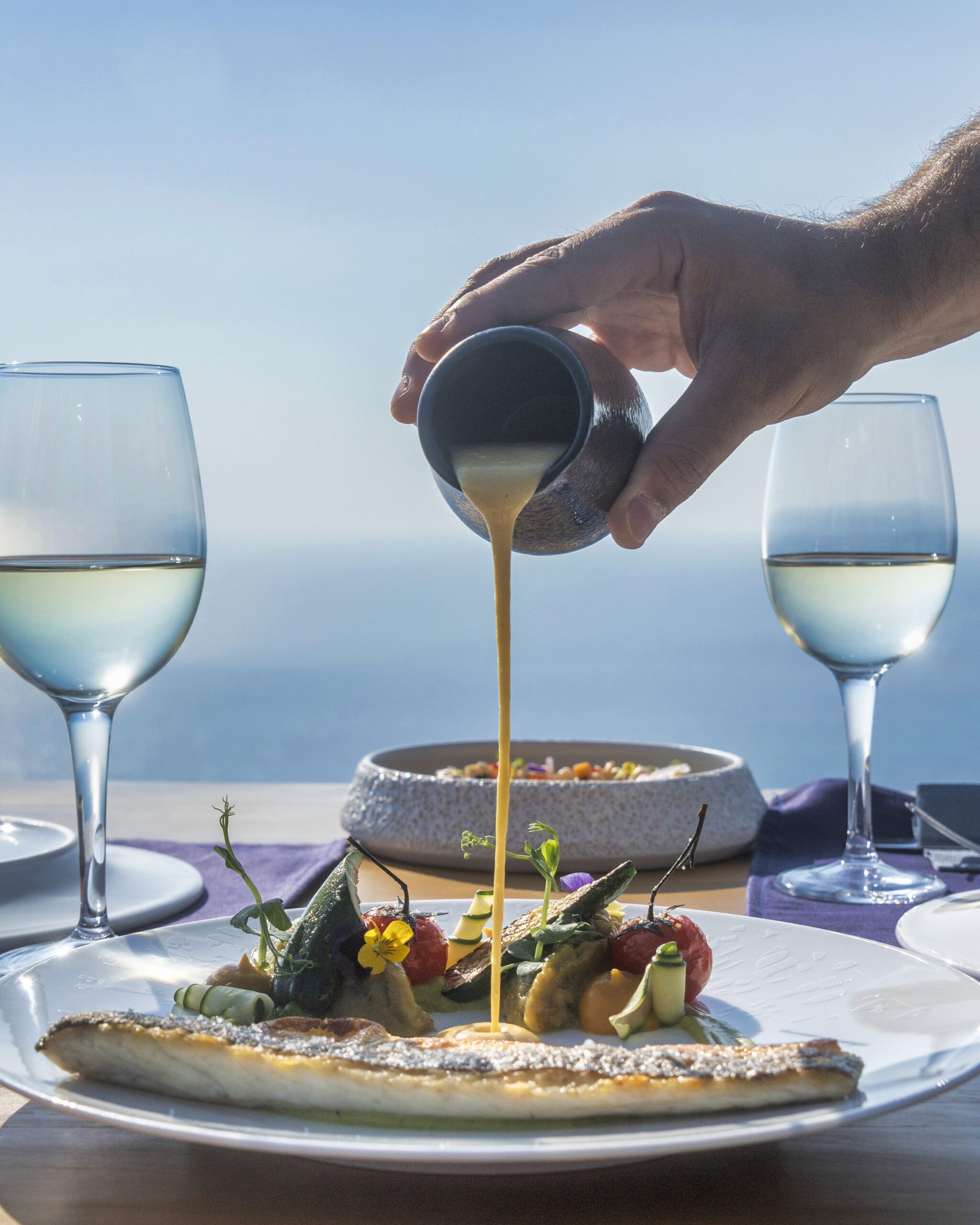 A hand pouring a creamy sauce over a dish with vegetables and fish, with two glasses of white wine on the table.