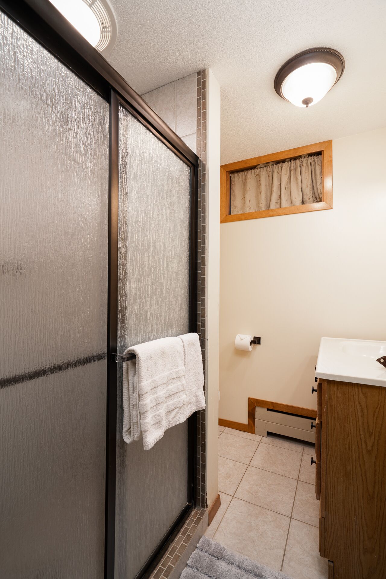 Ein Badezimmer mit Duschabtrennung aus Glas, einem an der Tür hängenden Handtuch, einem Holzkabinet und einem Fenster mit Vorhängen.