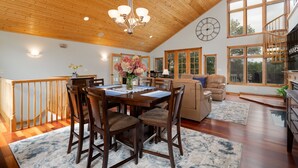 A spacious living area with a high wooden ceiling, a dining table with chairs, a sofa, and a large window offering a view of the outdoors.