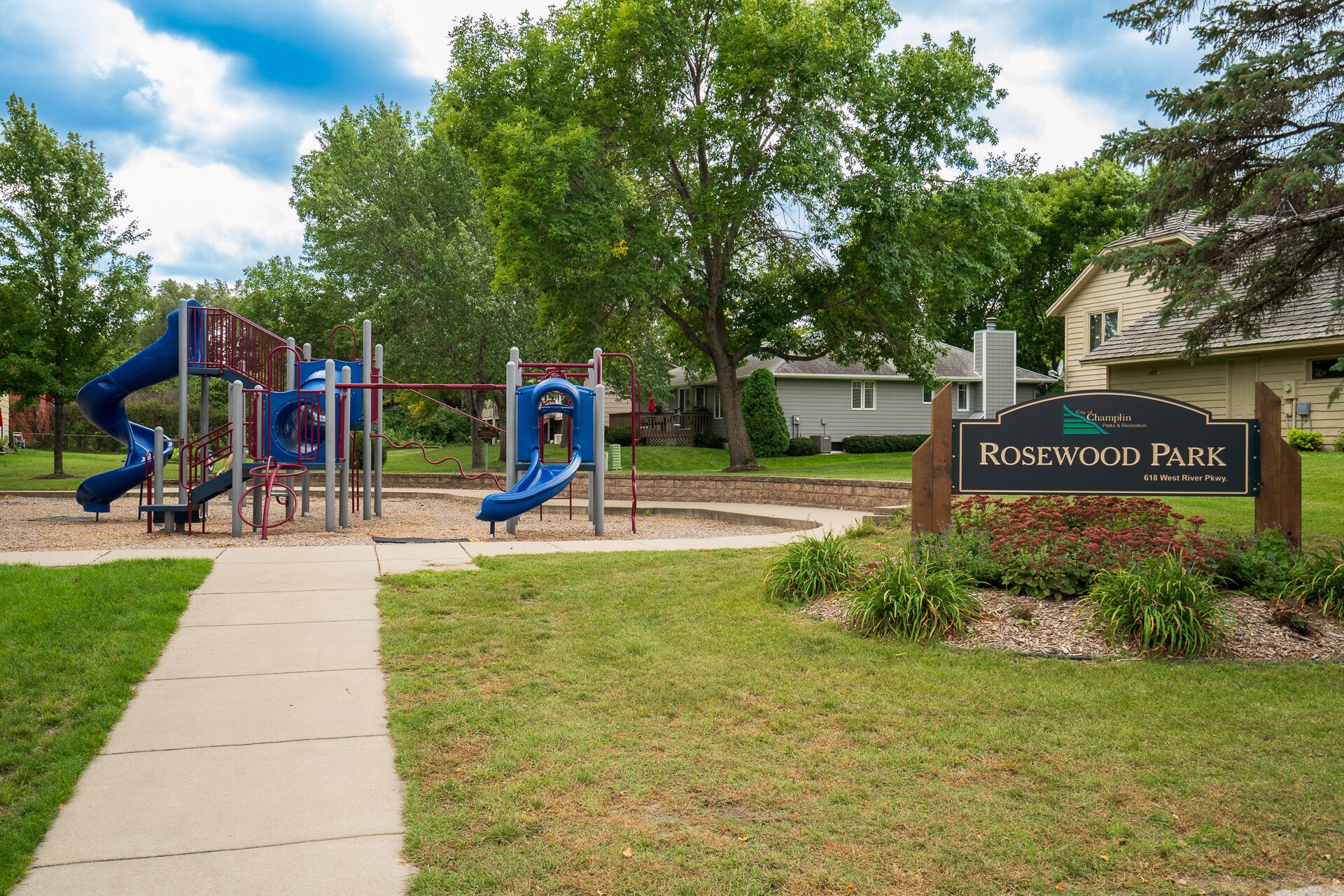 A playground with slides and climbing structures, surrounded by grass and trees. A sign indicates the name 'Rosewood Park'.