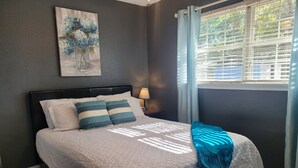 A bedroom with a bed, a nightstand, a lamp, a window with curtains, and a painting on the wall. - Great Gulf Breeze Getaway Near Pensacola Beach (Gulf Breeze)