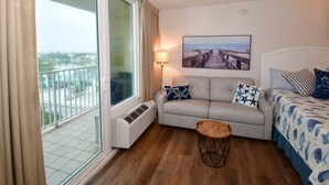A hotel room with a balcony, a sofa, a bed with patterned bedding, a small wooden table, and a wall-mounted lamp. - Beautiful Beach View Condo with heated pool. No Resort Fees. (Treasure Island)