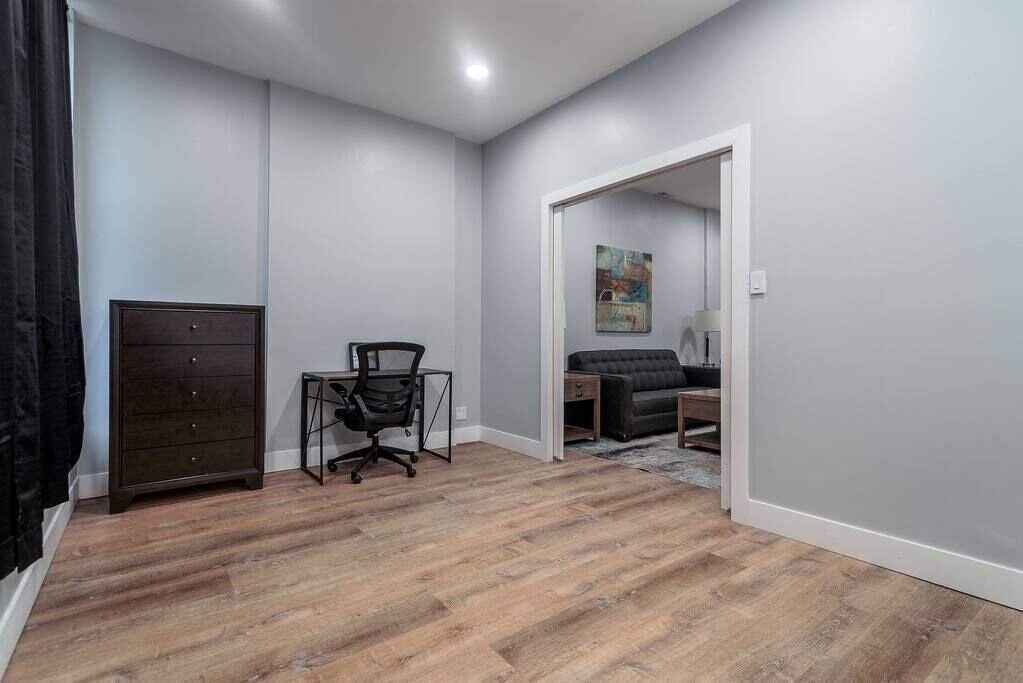 A room with a wooden floor, a dark-colored dresser, a black office chair, a gray sofa, and a painting on the wall.