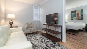 Ein modernes Wohnzimmer mit einem weißen Sofa, einem gemusterten Sessel, einem Fernseher auf einem Metallständer und einem Bett mit weißer Bettwäsche.