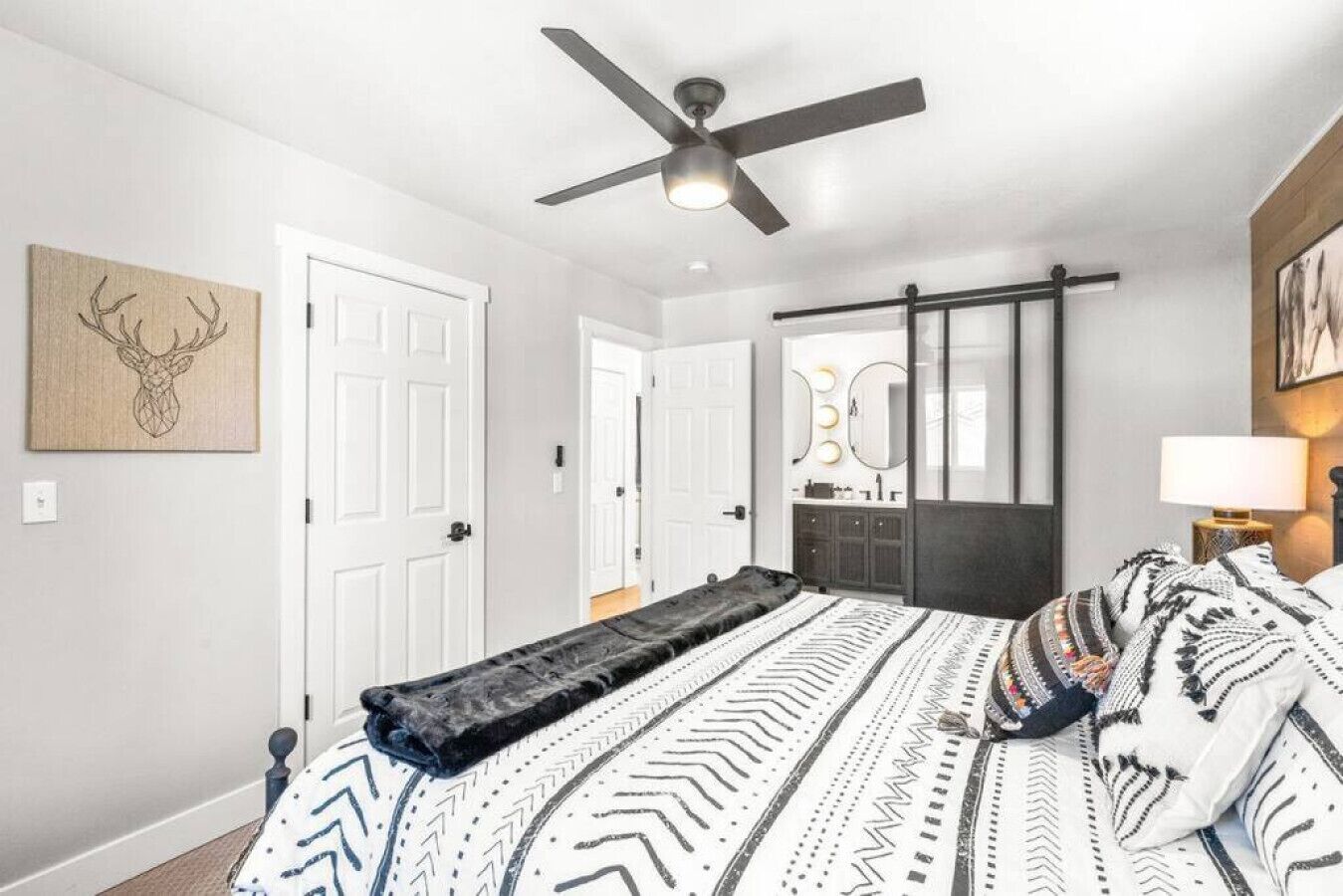 A bedroom with a bed, a ceiling fan, a door, a mirror, and a wall-mounted artwork.
