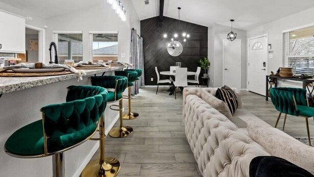 A modern kitchen with a dining area, featuring a bar with green velvet chairs, a tufted sofa, and a black accent wall.