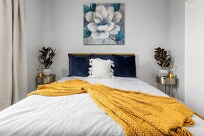 Un lit bien fait, recouvert d’une couverture blanche et jaune, avec un oreiller bleu foncé et un oreiller blanc orné d’un pompon blanc. Un tableau représentant une fleur est accroché au mur, au-dessus du lit.