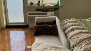 A bedroom with a sofa, a fluffy rug, a window with blinds, a radiator, and a view of buildings. - Stylish studio apartment in the heart of East Harlem (New York)