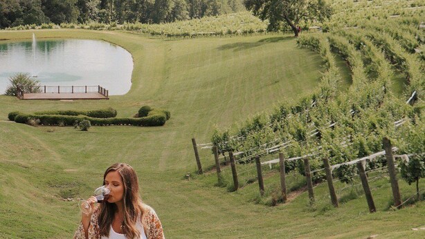 Una donna che cammina in un vigneto con un lago e un recinto di legno sullo sfondo.