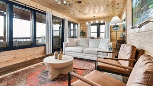 Un interior de cabaña acogedor, con un sofá, sillones, una mesita redonda y un ventanal grande.