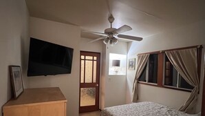Une chambre à coucher comprenant un lit, un ventilateur de plafond, un téléviseur fixé au mur, une armoire en bois et une porte donnant sur une autre pièce.