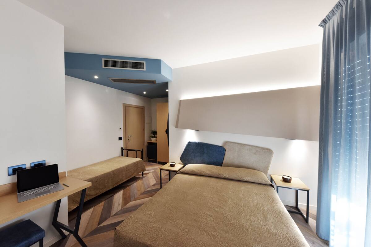A hotel room with a bed, a desk, a laptop, a chair, and a window with curtains.