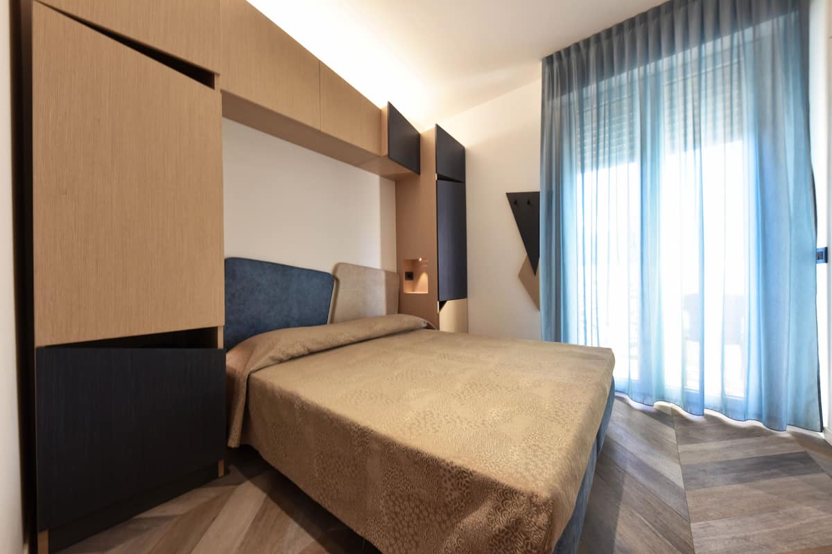 A modern hotel room with a bed, built-in wardrobe, and a large window with curtains.