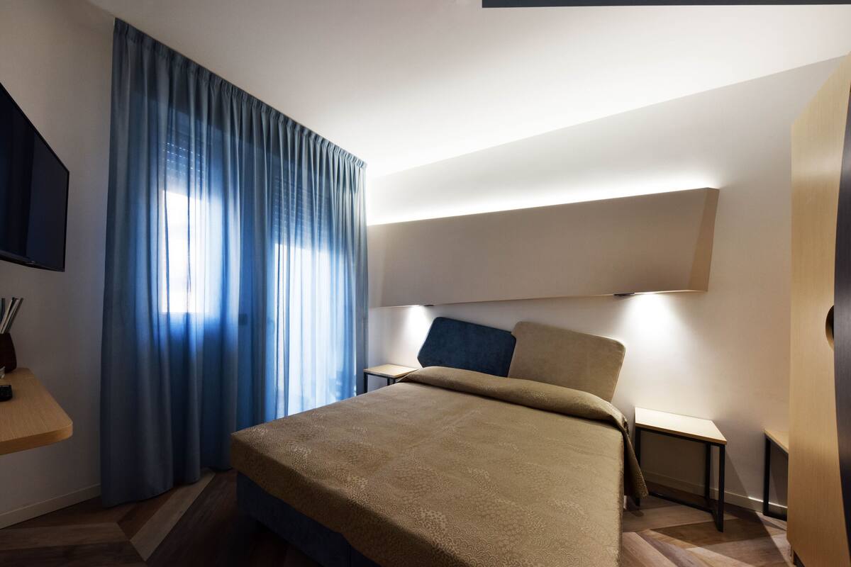 A hotel room with a bed, a bedside table, a TV, and a large window with curtains.