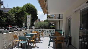 An outdoor cafe with tables and chairs, a decorative fence, and a view of parked cars and buildings in the background.