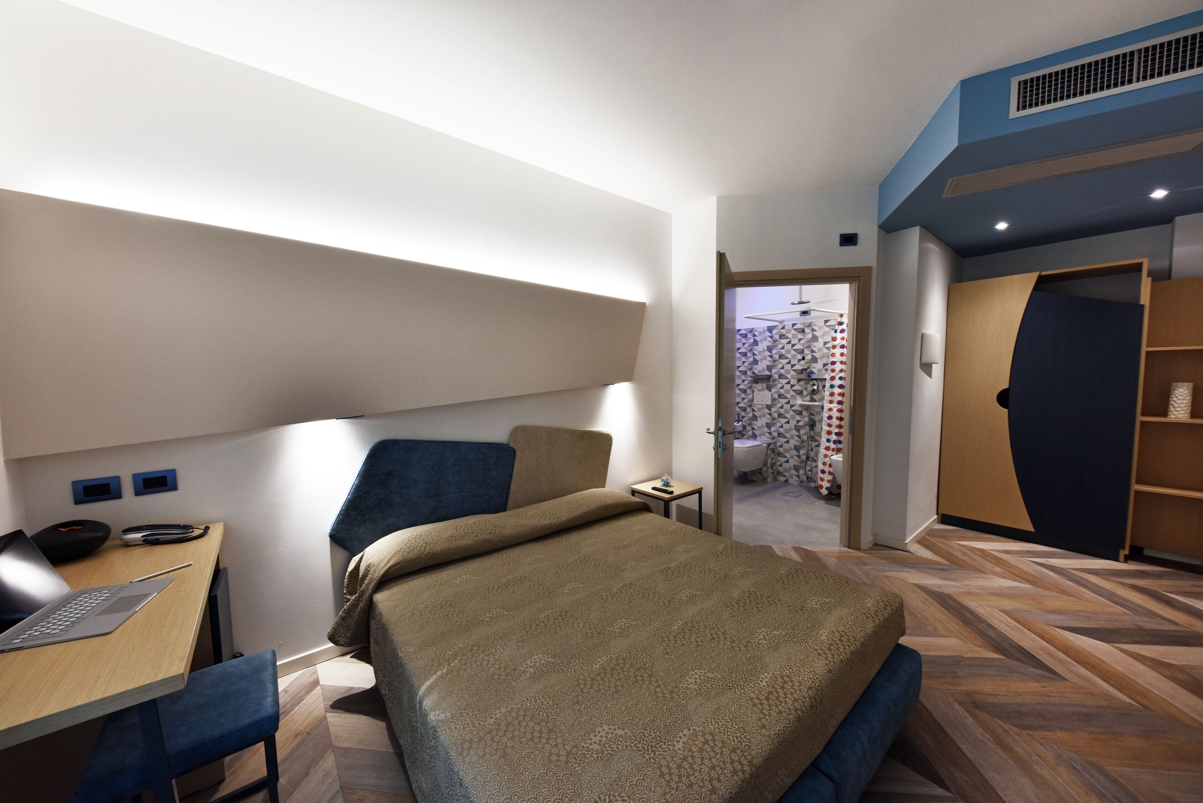 A modern hotel room with a bed, desk, and chair, a bathroom with a shower and toilet, and a small sitting area.