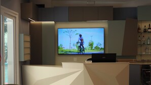 A modern reception area with a large TV screen displaying a cyclist, a sleek reception desk, and a bar area with various bottles.