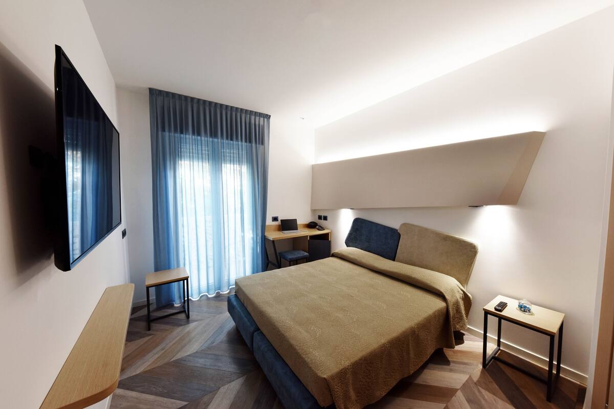 A modern hotel room with a large bed, a desk, a chair, a TV, and a small table.