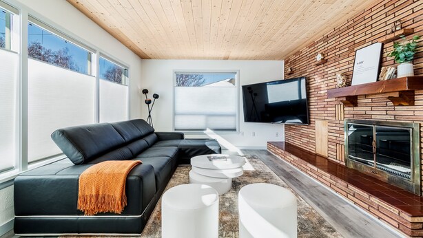 Een moderne woonkamer met een zwarte leren bank, witte ronde poufs, een flatscreen televisie en een open haard met bakstenen.