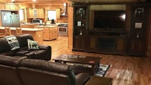 A spacious living room with a leather sofa, a coffee table, and a small wooden table. A kitchen with wooden cabinets and appliances is visible in the background. - 4500 sq ft Secluded Log Cabin Home on 17 acres (Waco)