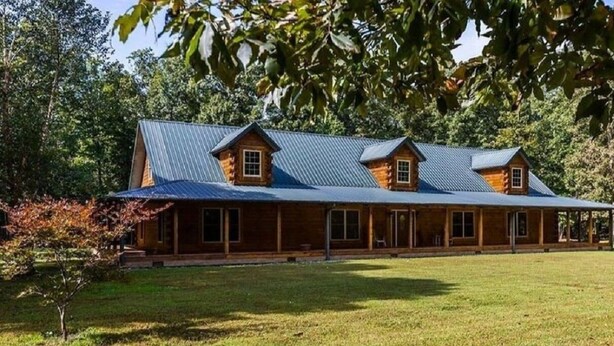 A large, two-story log cabin with a metal roof, surrounded by a well-maintained lawn and dense trees. - 4500 sq ft Secluded Log Cabin Home on 17 acres (Waco)