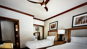 Habitación de hotel con una cama grande, una cama individual, un escritorio, una silla, una mesita de noche con lámpara, un ventilador de techo y un cuadro en la pared.