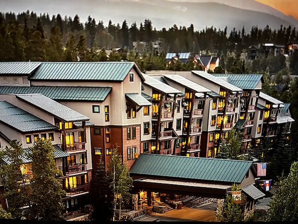 Um hotel com vários andares, telhado verde, diversas varandas e vista para uma cadeia montanhosa arborizada.