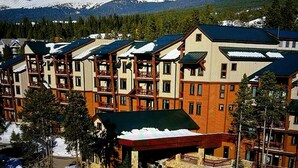 Un hotel de varios pisos con balcones, una zona de estacionamiento techada y montañas nevadas al fondo.