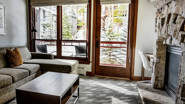 Una sala de estar con sofá, una mesa de centro y una chimenea. Se ven ventanas grandes que ofrecen vistas a árboles y edificios.