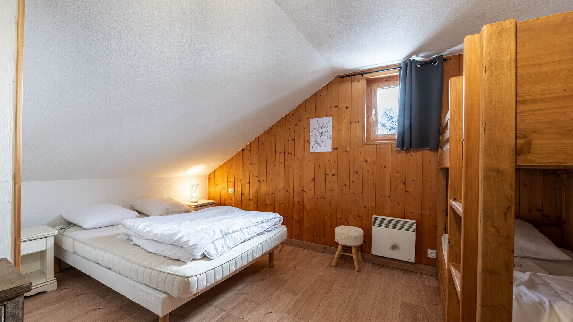 Un dormitorio acogedor en el ático con techo inclinado, paredes de madera, una cama individual, un taburete pequeño y un radiador.