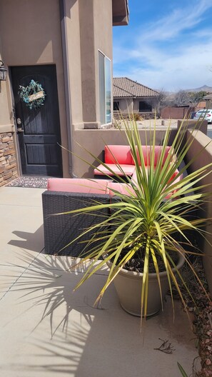 Relax in the serene outdoor seating area with a wicker chair and sofa, surrounded by lush greenery and a welcoming entrance.