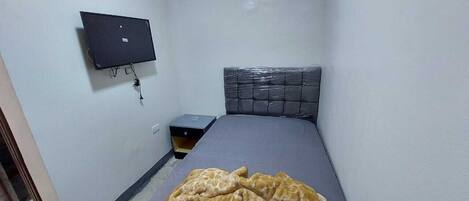 Una habitación pequeña y estrecha con una cama, una mesita de noche y un televisor montado en la pared.