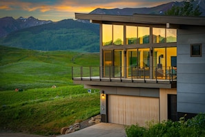 Un bâtiment moderne à deux étages, doté de grandes fenêtres, d’un balcon et offrant une vue sur un paysage montagneux.