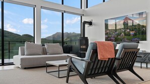 A modern living room with a sofa, two armchairs, a table, and a fireplace. Large windows offer a view of a mountainous landscape.