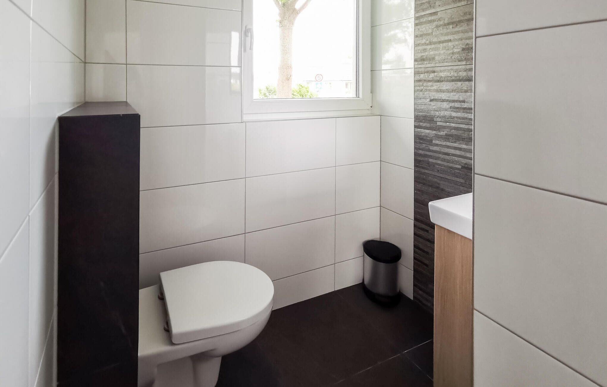Een moderne badkamer met witte tegels, een wit toilet, een zwarte prullenbak en een raam met uitzicht op bomen.