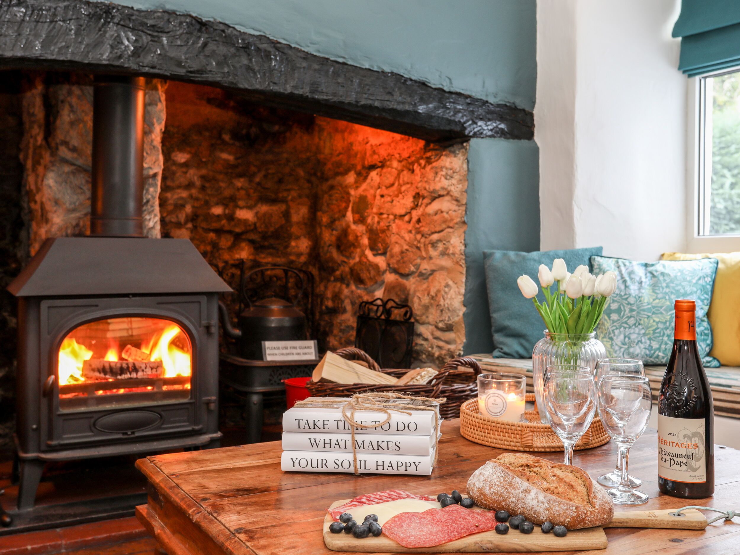Una stanza accogliente con camino in pietra, un tavolo di legno con libri, pane e formaggio, e una bottiglia di vino.