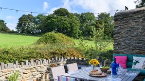 An outdoor patio with a stone wall, a table set for dining, and a stone wall with a view of a green field. - Ty Bach - Beaumaris (Beaumaris)
