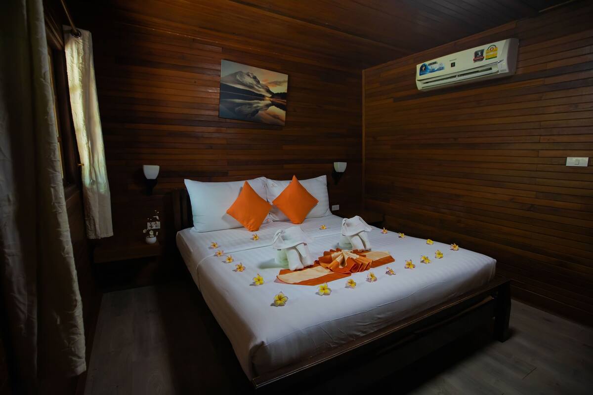 Una habitación con paneles de madera, una cama, almohadas, un cuadro, un equipo de aire acondicionado y una mesita de noche con una lámpara.
