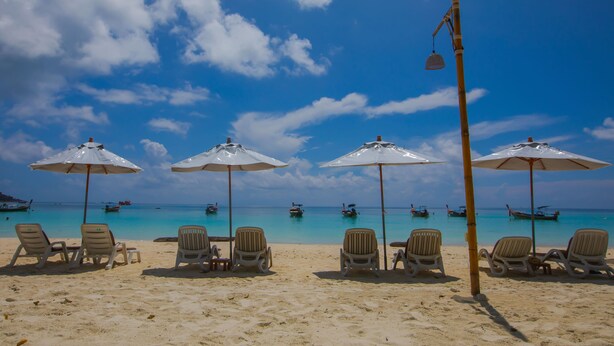 Une plage avec des chaises longues et des parasols, des bateaux sur l’eau et un ciel dégagé.