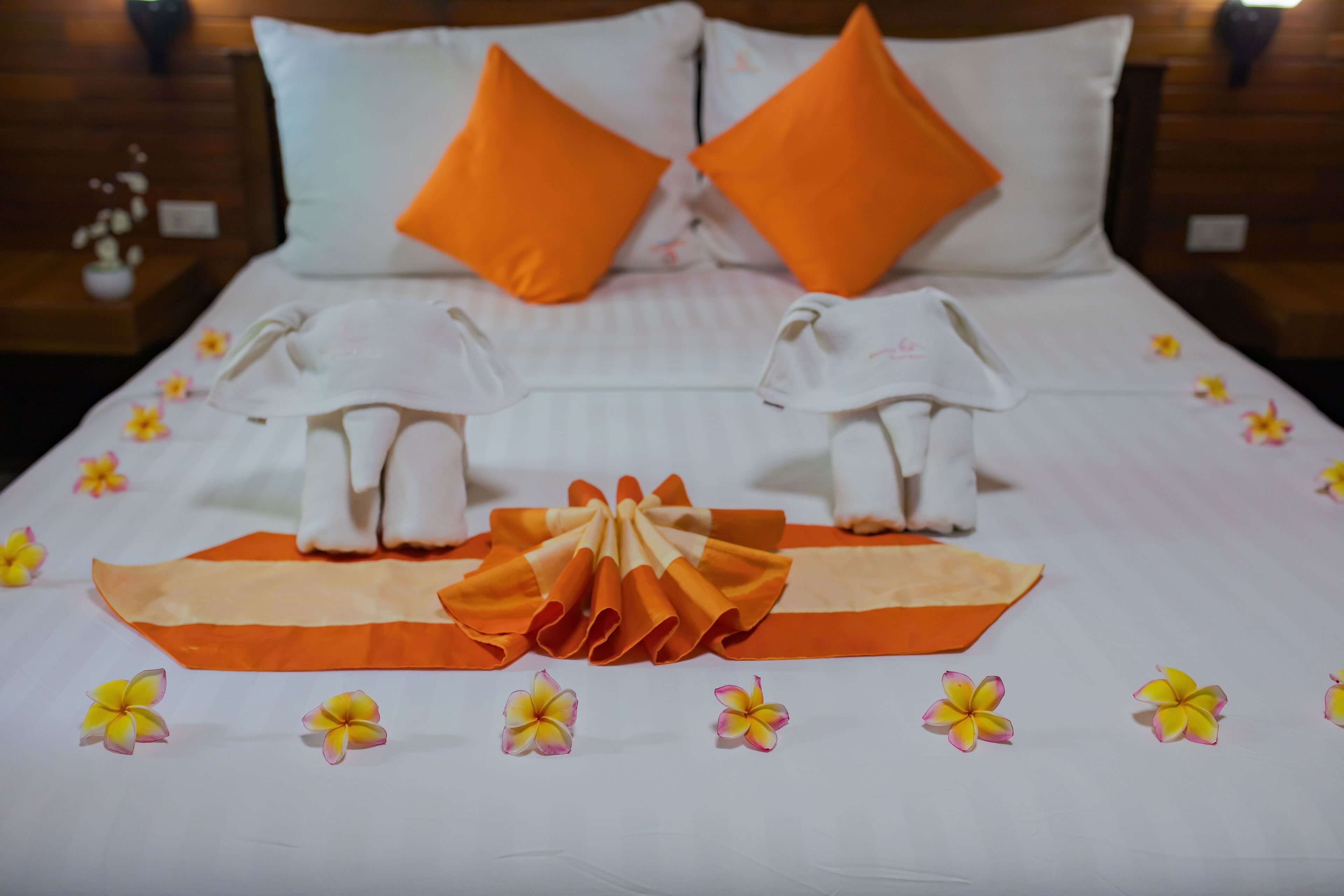 A neatly made bed with orange pillows, a folded towel, and a decorative bow.