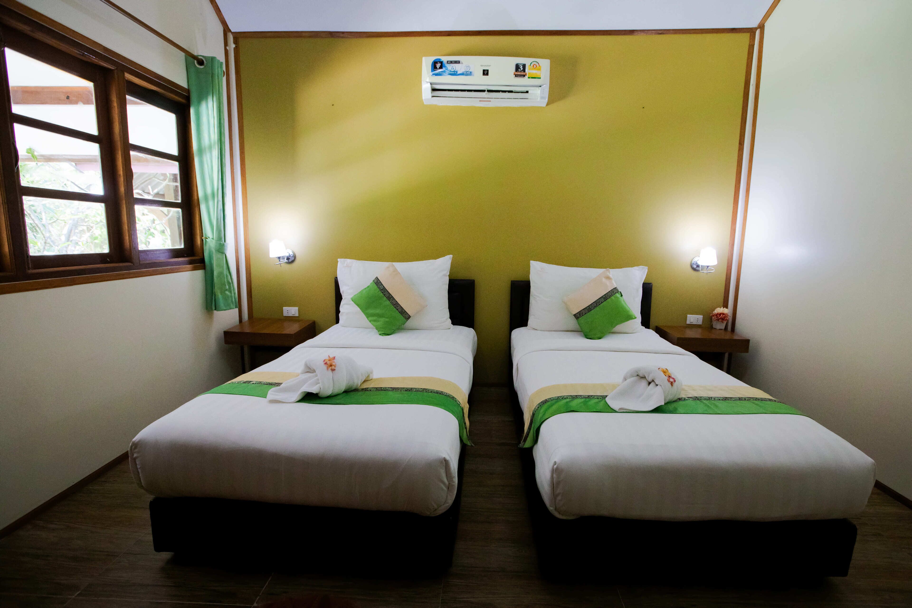 A hotel room with two beds, each with white linens, green pillows, and a green bedspread. There are nightstands with lamps, a wall-mounted air conditioner, and a window with a green curtain.