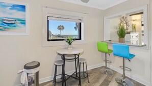 A small dining area with a round table, two stools, and a bar counter with two stools. There is a window with a view of palm trees and a boat.