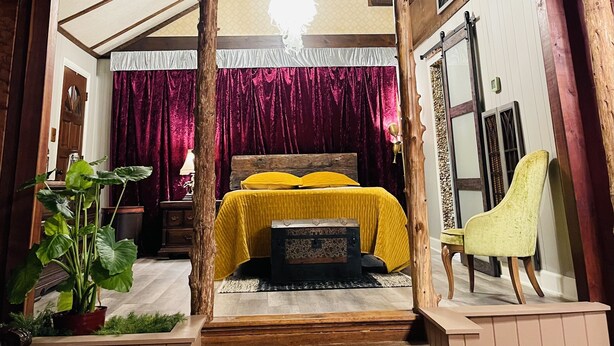 Une chambre à coucher rustique avec un plafond en bois, un lit recouvert d’une courtepointe jaune, une chaise en bois et une fenêtre ornée d’un cadre décoratif.