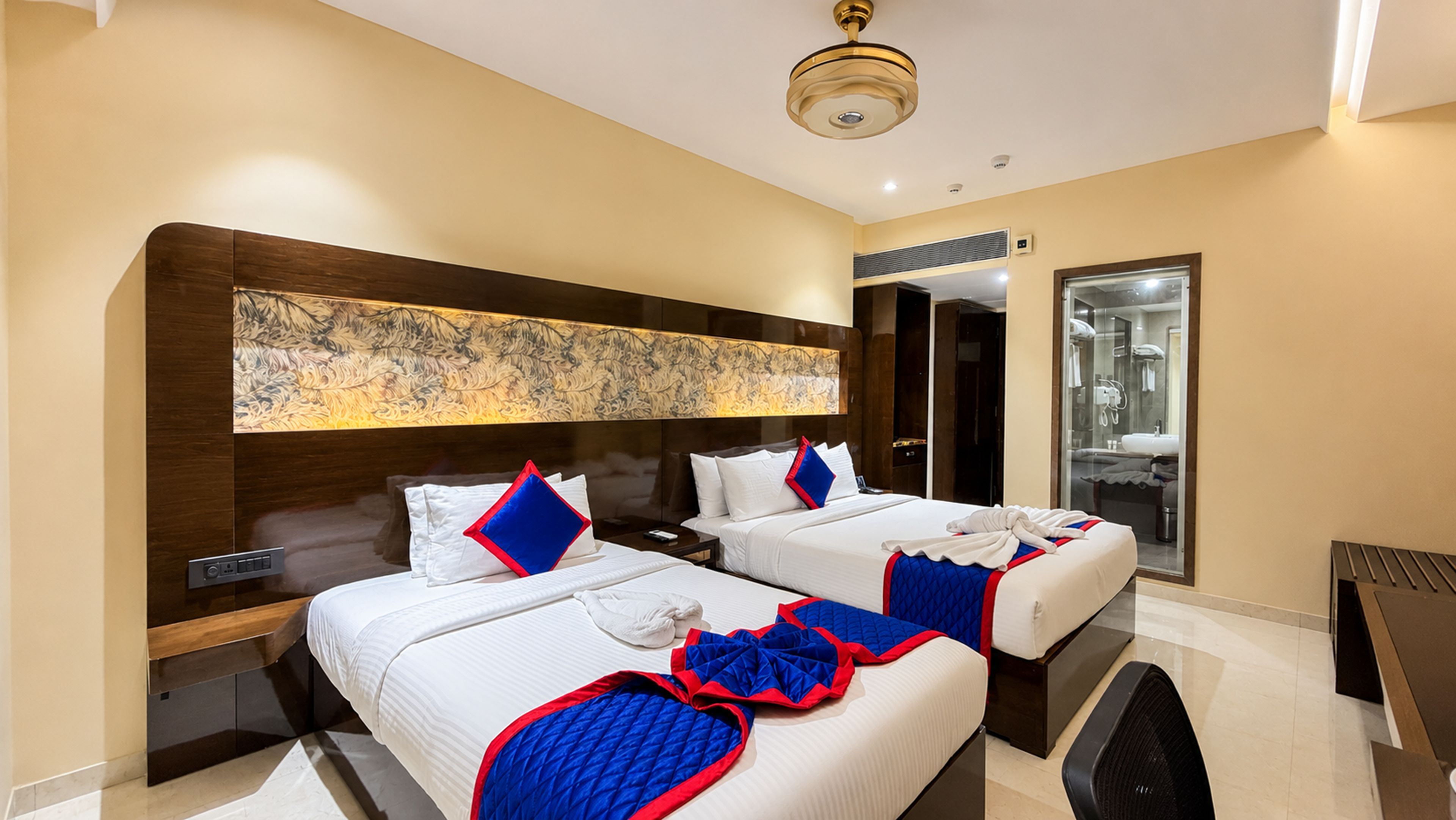 Habitación de hotel con dos camas, una obra de arte grande sobre la cabecera, un baño con mampara de vidrio y un ventilador de techo.