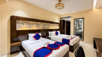 Habitación de hotel con dos camas, una obra de arte grande sobre la cabecera, un baño con mampara de vidrio y un ventilador de techo.