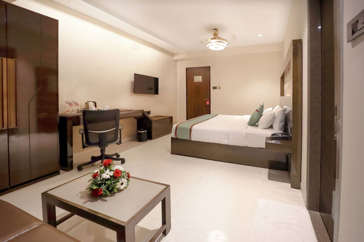 Quarto de hotel com cama, escrivaninha com cadeira, uma pequena mesa com arranjo de flores e televisão na parede.